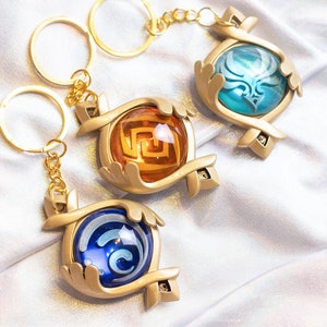 May include: Three gold keychains with different colored glass orbs. Each orb has a different design inside. The keychains are on a white fabric background.