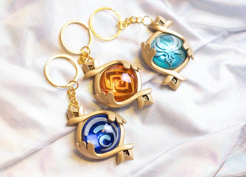 May include: Three gold keychains with different colored glass orbs. Each orb has a different design inside. The keychains are on a white fabric background.