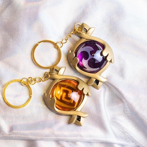 May include: Two gold keychains with purple and orange orbs. The orbs have a geometric design inside. The keychains are on a white fabric background.