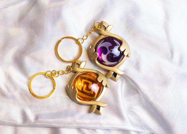 May include: Two gold keychains with purple and orange orbs. The orbs have a geometric design inside. The keychains are on a white fabric background.