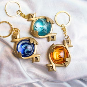 May include: Three gold keychains with different colored glass orbs. Each orb has a different design inside. The keychains are on a silver background.