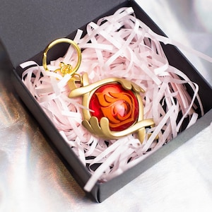 May include: A gold-colored keychain with a red and orange gem-like charm. The charm features a stylized flame design. The keychain is in a black box with pink tissue paper.