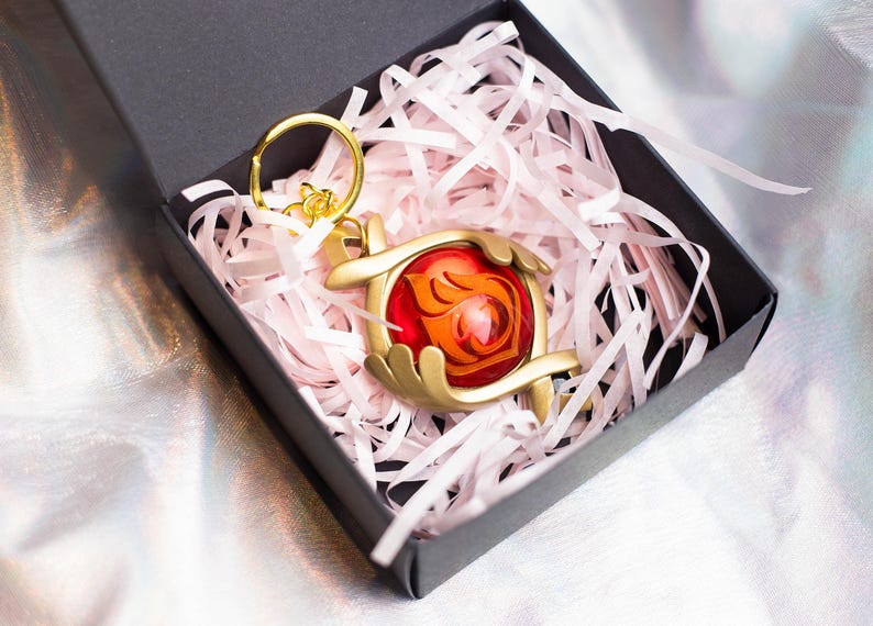 May include: A gold-colored keychain with a red and orange gem-like charm. The charm features a stylized flame design. The keychain is in a black box with pink tissue paper.