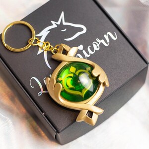 May include: Gold-colored key chain with a green gem in the center. The key chain is shaped like a pair of hands holding the gem. The key chain is on a black box with the word "Unicorn" printed on it.