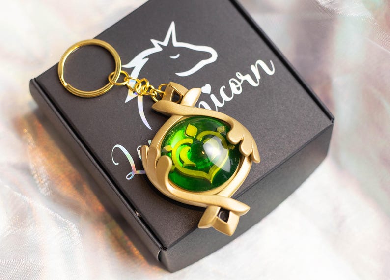May include: Gold-colored key chain with a green gem in the center. The key chain is shaped like a pair of hands holding the gem. The key chain is on a black box with the word "Unicorn" printed on it.