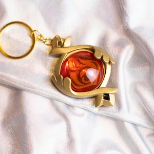 May include: A gold-colored keychain with a red and orange gem-like charm. The charm is shaped like a stylized heart with intricate details.