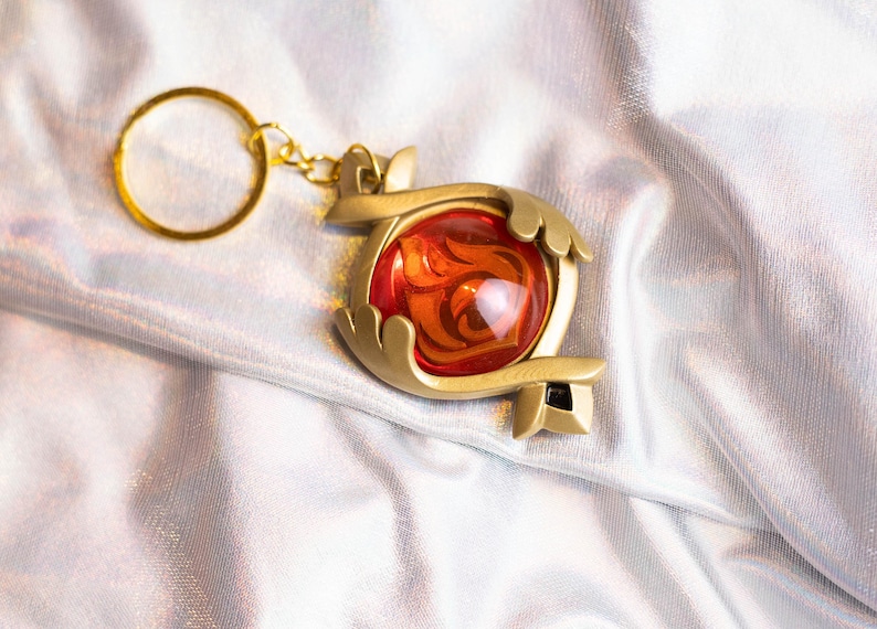 May include: A gold-colored keychain with a red and orange gem-like charm. The charm is shaped like a stylized heart with intricate details.