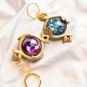 May include: Two gold keychains with ornate designs. One keychain features a blue gem with a snowflake design, the other keychain features a purple gem with a swirling design.