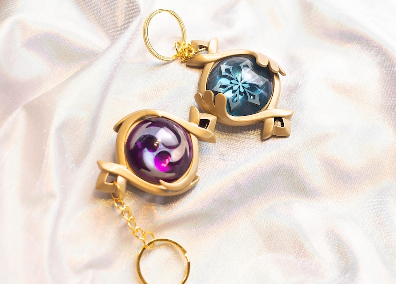 May include: Two gold keychains with ornate designs. One keychain features a blue gem with a snowflake design, the other keychain features a purple gem with a swirling design.