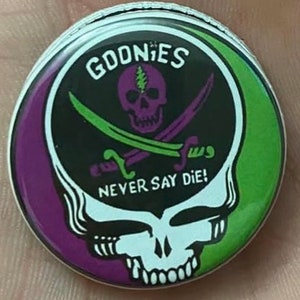 May include: A purple, green, and white button with a skull and crossbones graphic and the text "Goonies Never Say Die!"