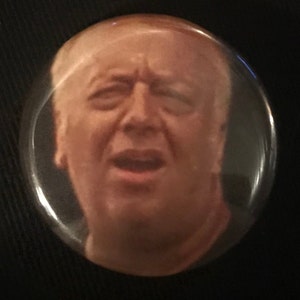 May include: A close-up of a button featuring a man's face with a surprised expression. The button is round and has a silver border.