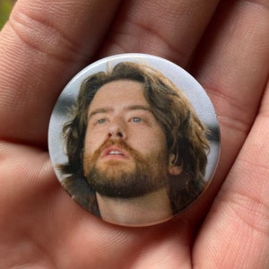 May include: A close-up of a button featuring a man with brown hair and a beard looking up. The button is white with a glossy finish.