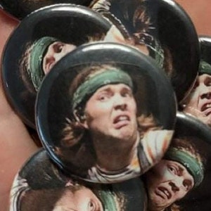 May include: A set of round buttons featuring a close-up image of a person with long brown hair wearing a green bandana and a colorful shirt. The person is looking directly at the camera with a surprised expression.
