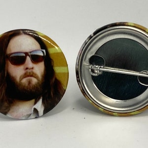 May include: A round button with a portrait of a man with long brown hair and a beard wearing sunglasses. The background is yellow and brown stripes.