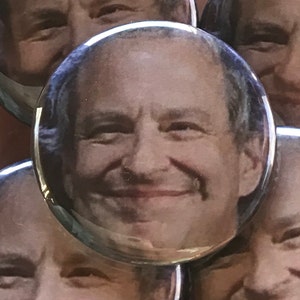 May include: A close-up of a man's face with a smiling expression. The image is printed on a round button.