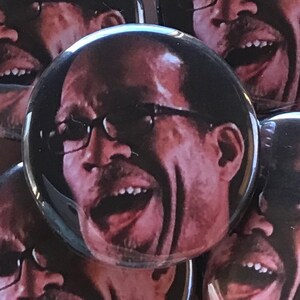 May include: A close-up of a button featuring a man with glasses and a wide open mouth, laughing. The button is black and has a glossy finish.