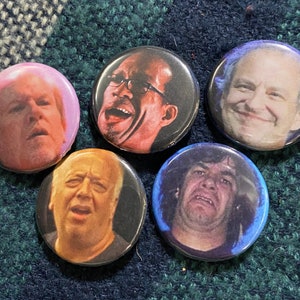 May include: Five round button pins with different images of people's faces. The buttons are in various colors, including pink, blue, and black.