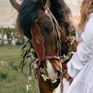 May include: A brown horse with a leather bridle and a floral garland around its neck. The horse is standing in a field of green grass.
