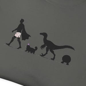 May include: A charcoal grey t-shirt featuring a black silhouette graphic of a person wearing a cape and polka-dot boxers, followed by a small cat with a purple bow, a velociraptor, and a round object, all walking in a line on a diagonal path.