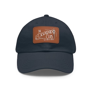 Op de afbeelding: Een marineblauwe baseballpet met een rechthoekige bruine leren patch aan de voorzijde. De patch bevat de tekst "THE DESPERADO CLUB" en "SO FUN IT HURTS" in witte letters, vergezeld van een grafische illustratie van een verticale dolk.