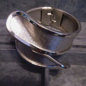 May include: A silver metal bangle bracelet with a curved, textured design. The bracelet has a hinged clasp.