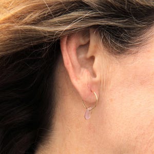 May include: A thin gold hoop earring featuring a small, smooth, translucent light pink teardrop-shaped charm suspended from the bottom. The earring is worn in the earlobe, showcasing the delicate metal wire and the soft pastel color of the pendant.