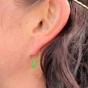 May include: A gold hoop earring featuring a single, smooth, translucent green sea glass charm. The small, circular gold wire hoop passes through the center of the rounded, pebble-shaped green glass piece, which hangs freely from the bottom of the hoop.