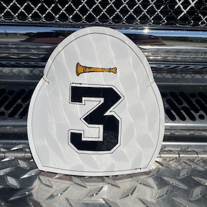 Pode incluir: Uma peça frontal para capacete de bombeiro em formato de escudo branco, apresentando um grande número 3 preto com contorno branco no centro. Acima do número, há um pequeno gráfico de uma trombeta dourada. A peça possui uma borda costurada.