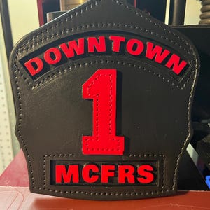 May include: A black leather firefighter helmet front shield with a shield-shaped design. It features the red text DOWNTOWN at the top, a large red number 1 in the center, and the red text MCFRS at the bottom, all stitched onto the black leather base.