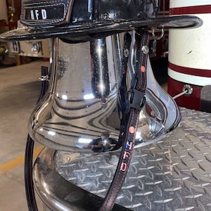 May include: A black firefighter helmet with a leather front piece displaying the number 3 and the letters HFD, resting on a polished chrome bell. The helmet features a black chin strap with red stitching and a metallic eagle ornament on top.