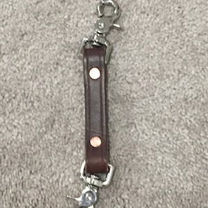 May include: A dark brown leather strap featuring two metallic rivets and silver-toned swivel snap hooks attached to both ends. The strap is rectangular in shape and serves as a connector or key fob accessory with a polished metal finish.