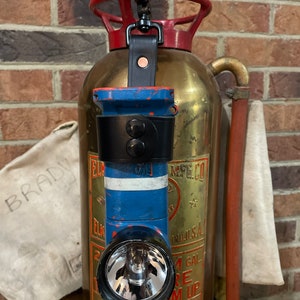 May include: A vintage brass fire extinguisher with a red handle and a brass body featuring text including ELKHART BRASS MFG. CO. A blue right-angle flashlight is attached to the top handle with a black leather strap and a metal swivel clip.