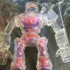 Może przedstawiać: Przezroczysty plastikowy robot-zabawka z tęczowym wzorem na głowie. Robot ma fioletowe i czerwone ciało z przezroczystą plastikową płytą piersiową. Robot stoi na białym tle.