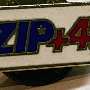 May include: A vintage ZIP+4 pin with a white background and the text "ZIP+4" in blue and red. The pin has a gold-coloured border and a star graphic. The pin is a collectible item.