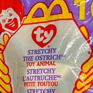 Pode incluir: Um saco de plástico roxo com um Ty Beanie Baby branco dentro. O saco tem o logotipo do McDonald's e as palavras "Stretchy the Ostrich" impressas nele.