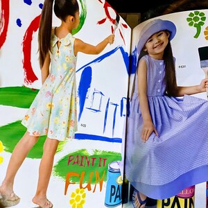 May include: Two young girls wearing dresses are painting on a white surface. The girl on the left is wearing a light blue and white floral dress and is painting a blue house. The girl on the right is wearing a light blue and white gingham dress and is holding a paintbrush. The text "Paint it Fun" is written in red and yellow letters on the surface. A blue paint can with the word "Paint" is visible in the lower right corner of the image.
