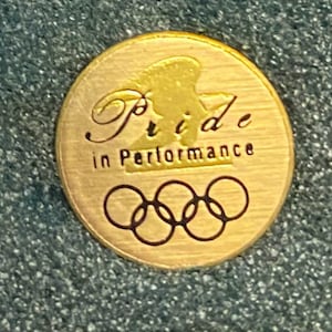 Puede incluir: Un pin redondo de color dorado con las palabras "Pride in Performance" y los anillos olímpicos. El pin presenta un diseño de águila estilizado. El pin está sobre un fondo texturizado color turquesa.