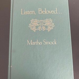 Puede incluir: Un libro de tapa dura verde con el título "Listen, Beloved..." y el nombre del autor "Martha Smock" en letras doradas. La portada presenta un diseño decorativo de dos motivos florales entrelazados.