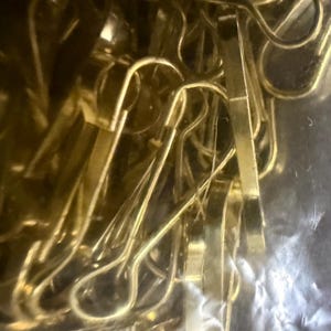 May include: Close-up of a collection of gold-colored metal paper clips. The clips have a classic design with a curved top and a straight, flat bottom. The clips are densely packed together, creating a shiny, reflective surface.