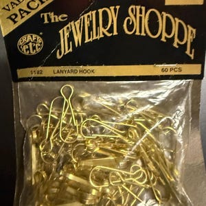 May include: A clear plastic bag containing gold-colored lanyard hooks. The package has a black and gold label that reads "The Jewelry Shoppe" and "Lanyard Hook". The package contains 60 pieces.