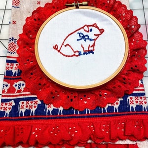 Może przedstawiać: Haftowany obręcz w kolorze czerwonym i białym z wzorem świni. Obręcz znajduje się na czerwono-niebieskim materiale z białym tłem.
