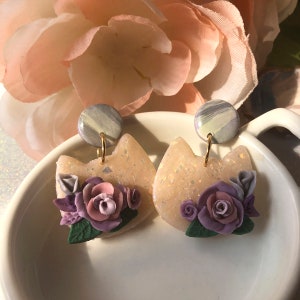 Puede incluir: Un par de pendientes en forma de gato de color rosa pastel con un diseño floral. Las flores están hechas de arcilla polimérica morada, rosa y blanca y tienen hojas verdes. Los pendientes tienen un fondo blanco y gris marmoleado.