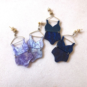 Puede incluir: Tres pares de pendientes con un diseño de traje de baño. Los pendientes están hechos de metal dorado y tienen un efecto marmoleado azul y morado.