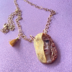 Puede incluir: Un collar de cadena dorada con un borla dorada y un colgante con el rostro de una mujer con un diseño dorado y rojo.