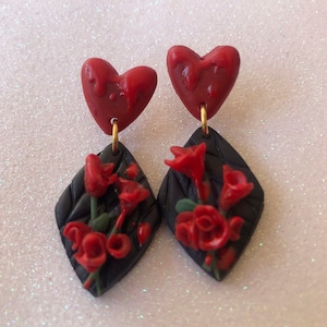 Puede incluir: Un par de pendientes colgantes con una base negra en forma de diamante y rosas rojas. La parte superior de cada pendiente es un corazón rojo.