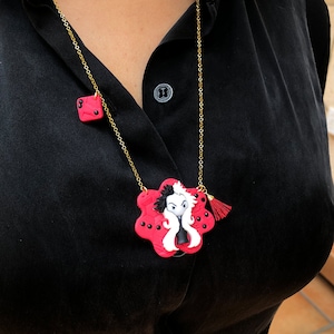 Puede incluir: Un collar de cadena dorada con un colgante rojo y negro con un personaje de dibujos animados. El colgante tiene forma de flor con un personaje blanco y negro en el centro. El collar también tiene un pequeño amuleto cuadrado rojo.