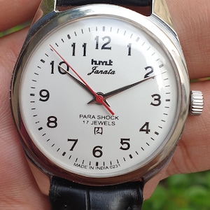 Może przedstawiać: Biały analogowy zegarek na rękę z czarnymi cyframi i wskazówkami. Tarcza zegarka zawiera napis "hmt Janata Para Shock 17 Jewels" i "Made in India 0231". Zegarek ma czarny skórzany pasek.