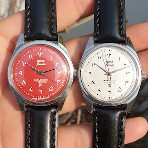 May include: Two wristwatches with black leather bands. The watch on the left has a red face with black Arabic numerals and the text "Humk Janata" and "Para Shock 17 Jewels". The watch on the right has a white face with black Arabic numerals and the text "Humk Janata" and "Para Shock 17 Jewels".