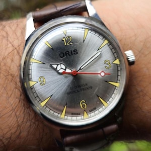 May include: A silver wristwatch with a white face and black hands. The watch has a brown leather strap and says "ORIS" and "17 JEWELS SHOCK PROOF" on the face.