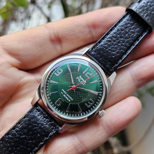 May include: A silver wristwatch with a green face and black leather band. The watch face has white numbers and a red second hand. The text on the watch face reads "Time Pilot Para Shock 17 Jewels".